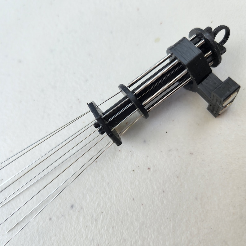 K&ouml;nnte beinhalten: Ein schwarzes Plastikwerkzeug mit mehreren d&uuml;nnen Metallnadeln, die sich von einem Ende erstrecken. Das Werkzeug hat eine Reihe von Federn und eine kleine Schlaufe am anderen Ende.