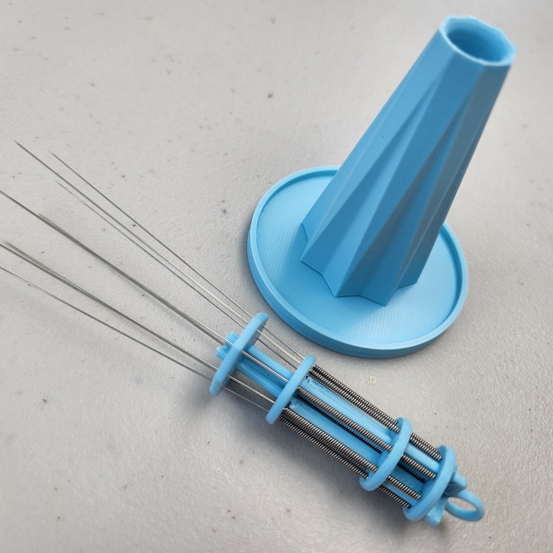 K&ouml;nnte beinhalten: Ein hellblaues Plastikwerkzeug mit mehreren d&uuml;nnen Metallnadeln, die an einem federbelasteten Mechanismus befestigt sind. Das Werkzeug ist zum Reinigen und Pflegen kleiner R&auml;ume konzipiert. Es wird mit einer passenden hellblauen Plastikbasis geliefert.