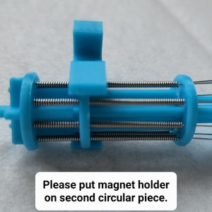 K&ouml;nnte beinhalten: Nahaufnahme eines blauen 3D-gedruckten Ger&auml;ts mit mehreren Metallfedern. Der Text "Bitte Magnet Halterung auf das zweite runde Teil setzen." ist unter dem Ger&auml;t sichtbar.