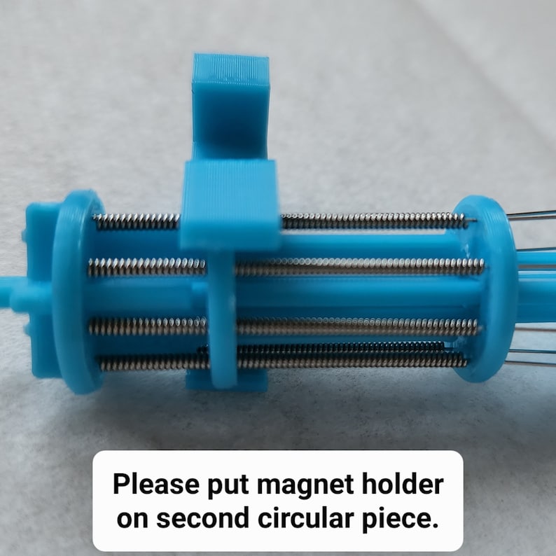 K&ouml;nnte beinhalten: Nahaufnahme eines blauen 3D-gedruckten Ger&auml;ts mit mehreren Metallfedern. Der Text "Bitte Magnet Halterung auf das zweite runde Teil setzen." ist unter dem Ger&auml;t sichtbar.