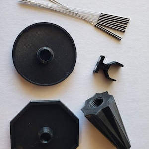 May include: A set of black 3D printed parts for a 3D printed pen. The set includes a round base, an octagonal base, a pointed tip, and a small black bracket. There are also 12 silver needles.