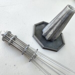 K&ouml;nnte beinhalten: Ein silbernes 3D-gedrucktes Modell eines Turms mit einem Sockel und einer dekorativen Spitze. Der Turm hat eine zylindrische Form mit einer Reihe von Ringen darum herum. Der Sockel ist sechseckig mit einer strukturierten Oberfl&auml;che. Die Spitze ist kegelf&ouml;rmig mit einer spitzen Spitze.