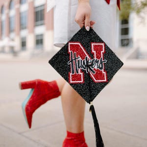May include: A black graduation cap with the word "Huskers" and a large red "N" in the center. The cap is held by a person wearing a white dress and red boots with blue soles. A red and white sash is also visible.