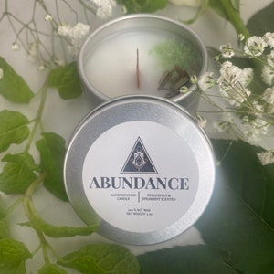 Puede incluir: Una vela de lata plateada con una parte superior de cera blanca y un cristal verde en su interior. La etiqueta dice "Abundance Manifestation Candle Eucalyptus & Spearmint Scented 100% Soy Wax Net Weight 2 oz."
