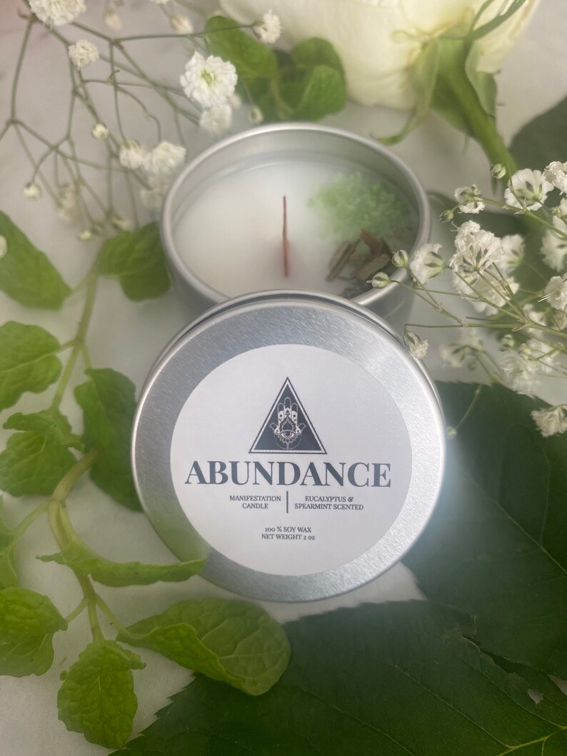 Puede incluir: Una vela de lata plateada con una parte superior de cera blanca y un cristal verde en su interior. La etiqueta dice "Abundance Manifestation Candle Eucalyptus & Spearmint Scented 100% Soy Wax Net Weight 2 oz."