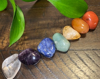 May include: Seven chakra stones arranged in a semi-circle on a wooden surface. The stones are clear, amethyst, blue, green, yellow, orange, and red.