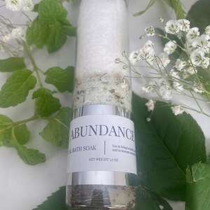 Puede incluir: Un frasco de vidrio plateado con una etiqueta blanca que dice "ABUNDANCE AL BATH SOAK" y "NET WEIGHT 3.7 OZ". El frasco est&aacute; lleno de un ba&ntilde;o de inmersi&oacute;n blanco y hierbas verdes. El frasco est&aacute; rodeado de flores blancas y hojas verdes.