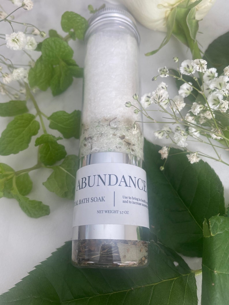 Puede incluir: Un frasco de vidrio plateado con una etiqueta blanca que dice "ABUNDANCE AL BATH SOAK" y "NET WEIGHT 3.7 OZ". El frasco est&aacute; lleno de un ba&ntilde;o de inmersi&oacute;n blanco y hierbas verdes. El frasco est&aacute; rodeado de flores blancas y hojas verdes.