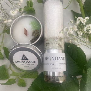 Puede incluir: Un tarro plateado con una etiqueta blanca que dice "ABUNDANCE RITUAL BATH SOAK" y "NET WEIGHT 3.7 OZ". El tarro est&aacute; lleno de sales de ba&ntilde;o blancas y hierbas verdes. El tarro est&aacute; sobre una superficie blanca con hojas verdes.