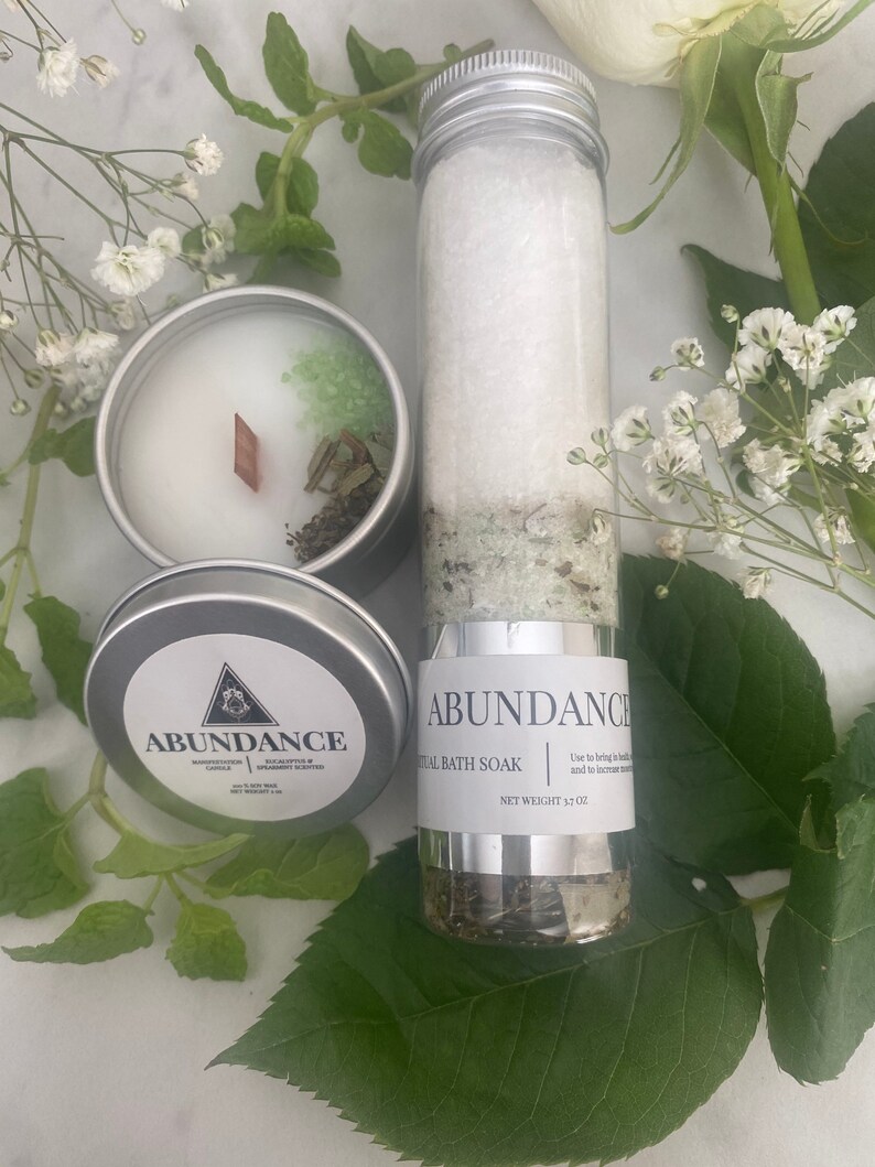 Puede incluir: Un tarro plateado con una etiqueta blanca que dice "ABUNDANCE RITUAL BATH SOAK" y "NET WEIGHT 3.7 OZ". El tarro est&aacute; lleno de sales de ba&ntilde;o blancas y hierbas verdes. El tarro est&aacute; sobre una superficie blanca con hojas verdes.