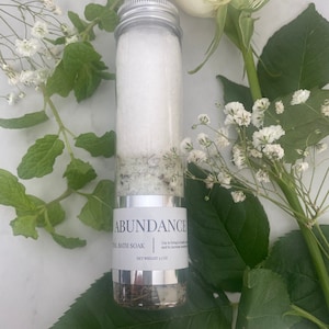 Puede incluir: Un tubo plateado lleno de sales de ba&ntilde;o blancas y etiquetado "Abundance Ritual Bath Soak" con un peso neto de 3,5 oz. El tubo est&aacute; rodeado de flores blancas y hojas verdes.