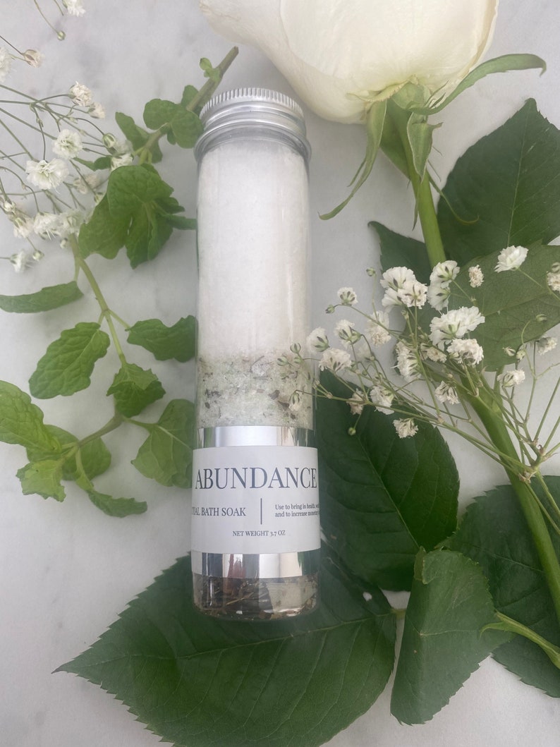 Puede incluir: Un tubo plateado lleno de sales de ba&ntilde;o blancas y etiquetado "Abundance Ritual Bath Soak" con un peso neto de 3,5 oz. El tubo est&aacute; rodeado de flores blancas y hojas verdes.