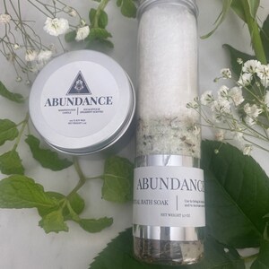 Puede incluir: Un tarro blanco con tapa plateada etiquetado como "Abundance Ritual Bath Soak" con un peso neto de 3,7 oz. El tarro est&aacute; lleno de un ba&ntilde;o de inmersi&oacute;n verde y blanco. El tarro est&aacute; sobre una superficie blanca con hojas verdes.