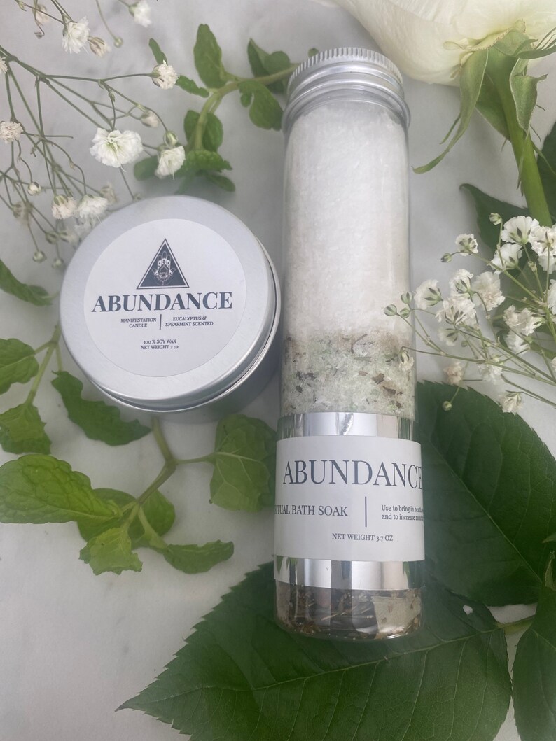 Puede incluir: Un tarro blanco con tapa plateada etiquetado como "Abundance Ritual Bath Soak" con un peso neto de 3,7 oz. El tarro est&aacute; lleno de un ba&ntilde;o de inmersi&oacute;n verde y blanco. El tarro est&aacute; sobre una superficie blanca con hojas verdes.