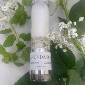 Puede incluir: Un tubo de vidrio transparente lleno de sales de ba&ntilde;o blancas y hierbas verdes. El tubo tiene una tapa plateada y una etiqueta blanca que dice "ABUNDANCE" y "TAL BATH SOAK". La etiqueta tambi&eacute;n incluye el texto "Usar para traer salud y aumentar el dinero" y "PESO NETO 3,7 OZ".