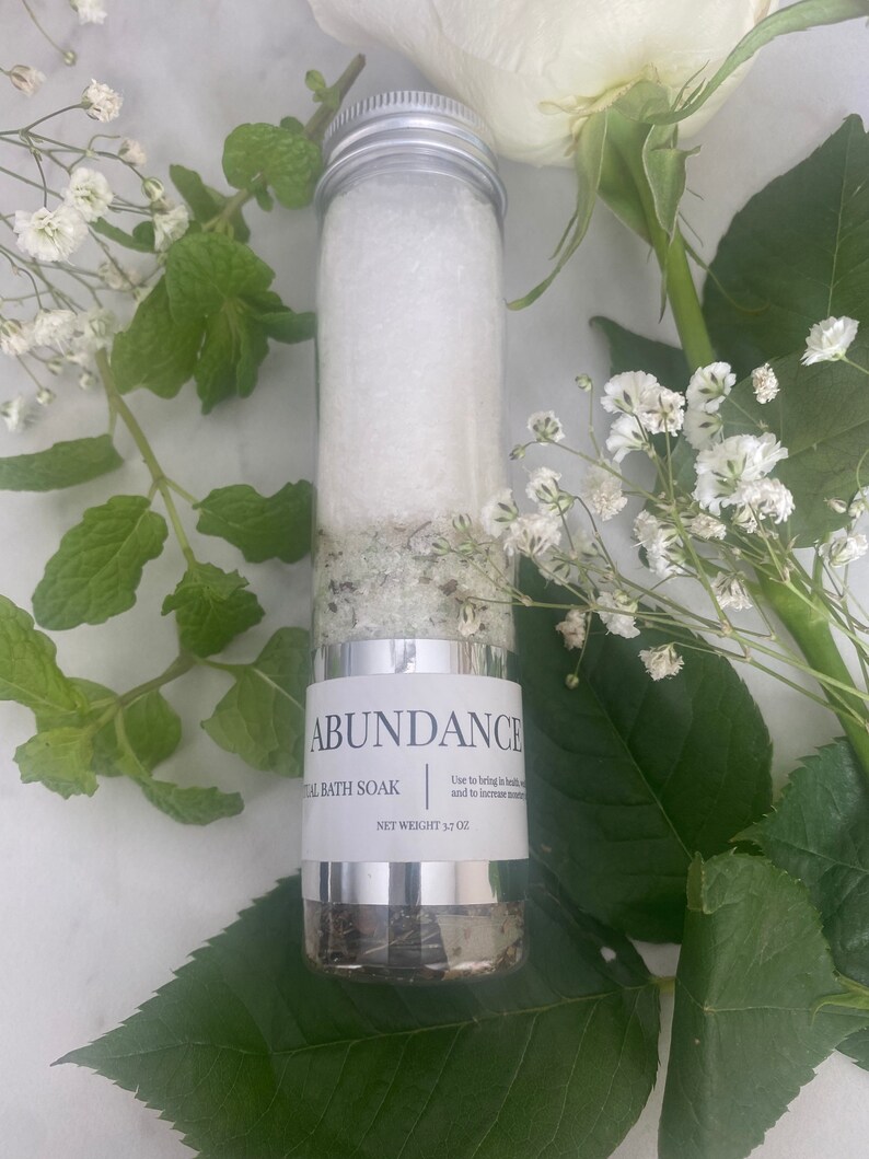 Puede incluir: Un tubo de vidrio transparente lleno de sales de ba&ntilde;o blancas y hierbas verdes. El tubo tiene una tapa plateada y una etiqueta blanca que dice "ABUNDANCE" y "TAL BATH SOAK". La etiqueta tambi&eacute;n incluye el texto "Usar para traer salud y aumentar el dinero" y "PESO NETO 3,7 OZ".