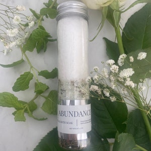 Puede incluir: Un tubo de vidrio transparente lleno de sales de ba&ntilde;o blancas y hierbas verdes. El tubo tiene una tapa plateada y una etiqueta que dice "Abundance Ritual Bath Soak" y "Peso neto 3,7 oz".