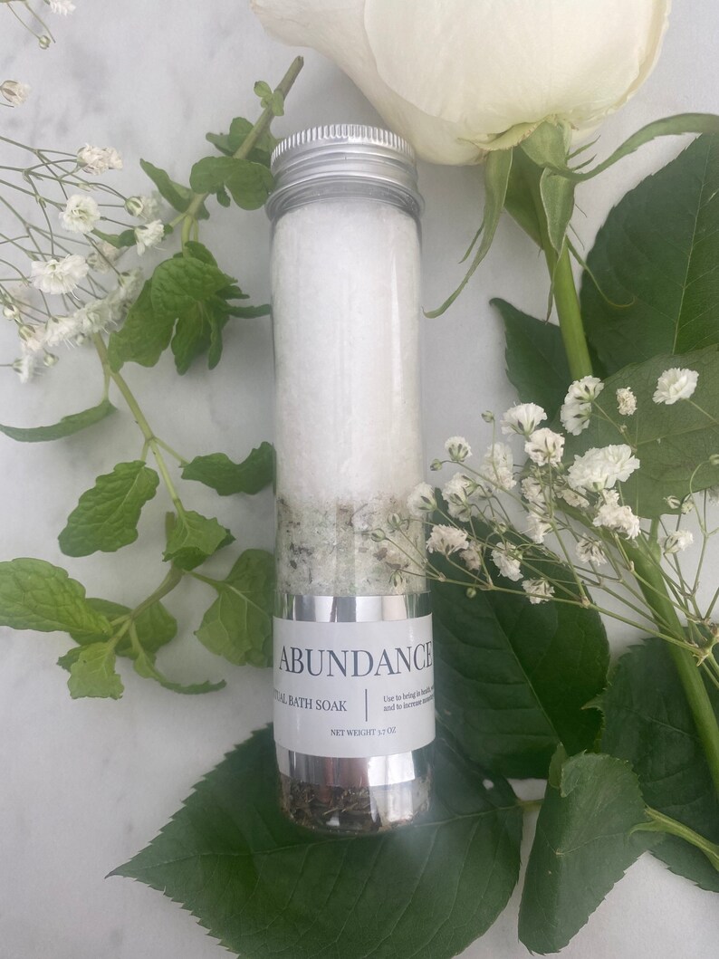 Puede incluir: Un tubo de vidrio transparente lleno de sales de ba&ntilde;o blancas y hierbas verdes. El tubo tiene una tapa plateada y una etiqueta que dice "Abundance Ritual Bath Soak" y "Peso neto 3,7 oz".