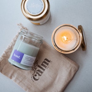 May include: Three glass jar candles with gold lids and a beige drawstring bag. One candle is lit, and another has a purple label that reads "bond." The bag has the text "little calm" and "EST. 21".