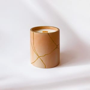 May include: A cylindrical candle in a peach-colored container with gold-colored crackle design. The candle has a wooden wick and a white wax fill. The candle is a decorative home decor item.