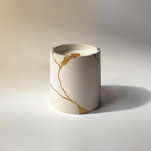 May include: A cylindrical, off-white candle in a concrete vessel, adorned with gold-colored crackle detailing. The candle has a wooden wick and is set against a white background, creating a minimalist aesthetic.