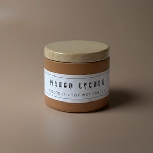 May include: A round, terracotta-colored candle jar with a wooden lid. The label reads "Mango Lychee Coconut + Soy Wax Candle" in a clean, sans-serif font. The candle sits on a neutral, light brown surface.