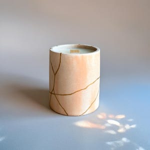 Peut inclure: Une bougie cylindrique dans un contenant pêche avec des lignes dorées craquelées. La bougie a une surface supérieure en cire blanche et une mèche en bois. Le contenant mesure environ 10 cm de haut et 7,6 cm de diamètre.