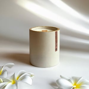 May include: An off-white cylindrical candle in a concrete holder with a wooden wick. The candle has a light brown rim and a label that reads "BIKO-HAUS". White flowers with yellow centres are in the foreground.