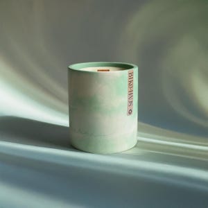 以下が含まれることがあります： 明るい緑と白の大理石模様の円筒形のキャンドル。キャンドルには木の芯があり、側面に「BIKO-HAUS」の文字が縦に印刷されています。キャンドルは水色の表面に置かれています。