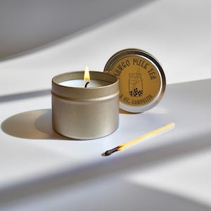 May include: A gold tin candle with a lit wick. The candle is labeled "Mango Milk Tea" and is 4 oz. The lid of the tin is off and sitting next to the candle. A used match is also next to the candle.