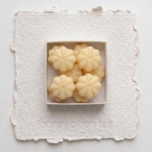 May include: A square box containing several pale yellow, flower-shaped wax melts. The box is centered on a piece of textured, deckle-edged off-white paper with the embossed text "honey citron tea" visible at the bottom of the paper.
