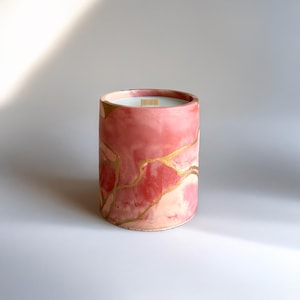 Peut inclure: Bougie cylindrique dans un contenant en céramique. Le contenant est peint avec un motif marbré rose et blanc, accentué de veines dorées. La bougie a une mèche en bois et est remplie de cire blanche.