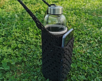 May include: A black crocheted bottle holder with a clear glass water bottle and a black phone inside. The holder is hanging from a black strap and is sitting on a bed of green grass.