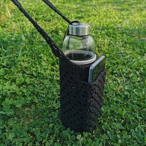 May include: A black crocheted bottle holder with a clear glass water bottle and a black phone inside. The holder is hanging from a black strap and is sitting on a bed of green grass.