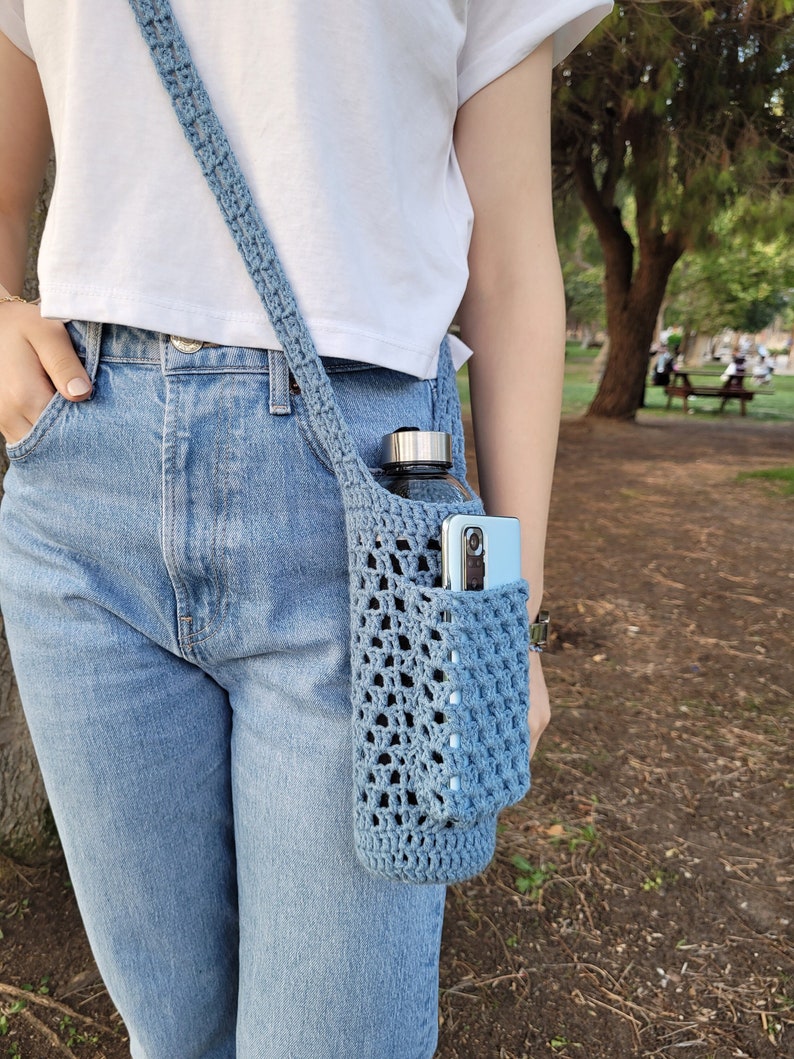 May include: A blue crocheted bag worn as a crossbody bag. The bag has a pocket for a water bottle and a phone. The bag is worn over a white t-shirt and blue jeans.