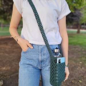 May include: A person wearing blue jeans and a white t-shirt is carrying a dark green crocheted bag with a water bottle and a phone inside.