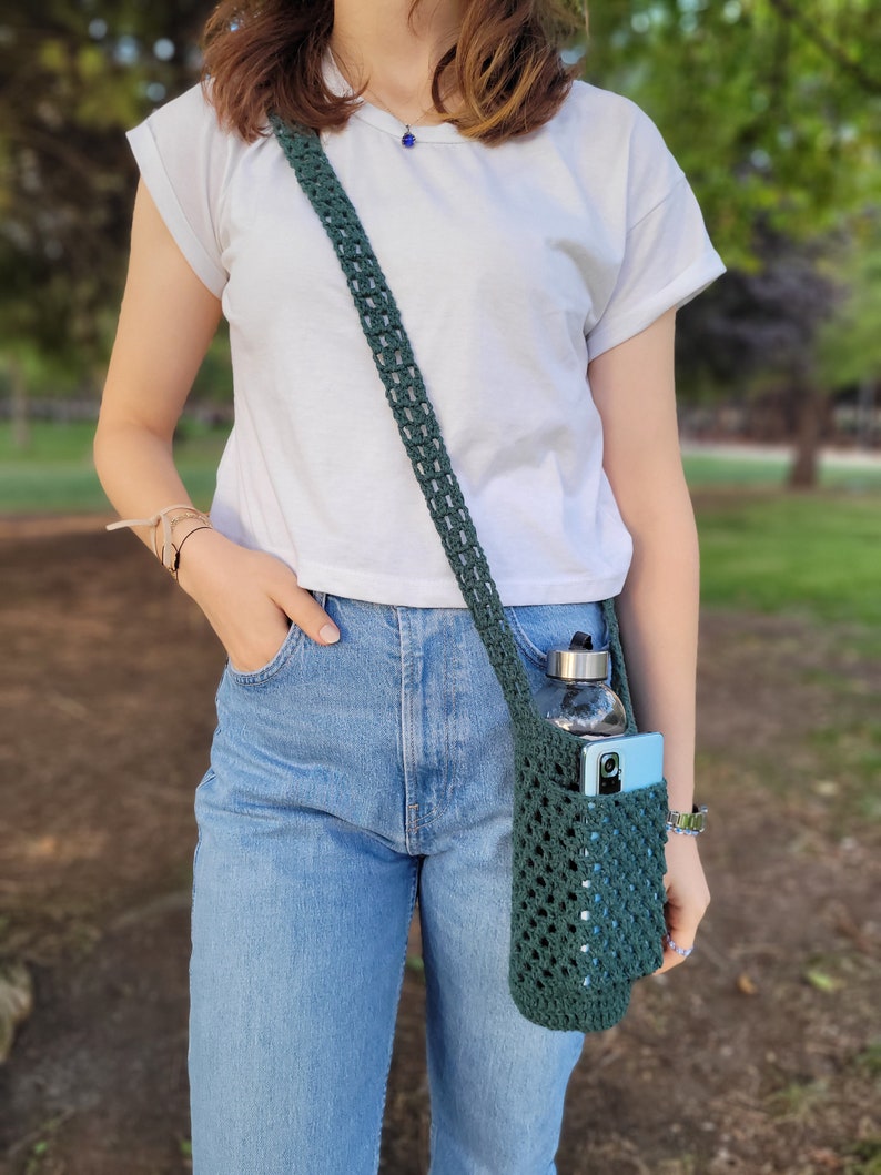 May include: A person wearing blue jeans and a white t-shirt is carrying a dark green crocheted bag with a water bottle and a phone inside.