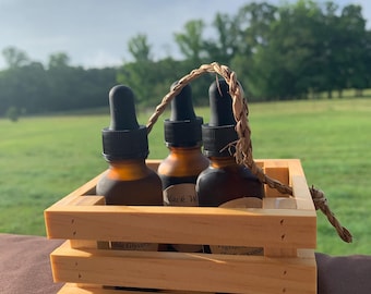 May include: Three amber glass dropper bottles with black caps are arranged in a small, light-colored wooden crate. The crate has a woven handle. Labels on the bottles read "Vegetable Glycerin" and "Black W". The background features a green field and trees.