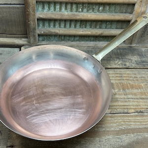 May include: A copper frying pan with a brass handle. The pan has a smooth, shiny surface and is sitting on a wooden surface.