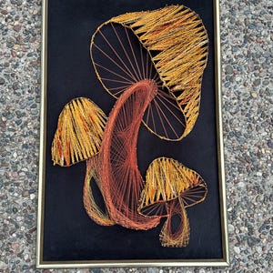 May include: A framed string art piece featuring three mushrooms in shades of orange and yellow against a black background. The artwork is encased in a gold-colored frame, creating a striking contrast.