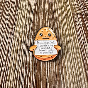 Puede incluir: Un pin de estilo caricatura con forma de patata sonriente con el texto "Positive potato" y un mensaje motivador. El pin es de color naranja y blanco.