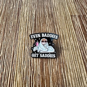 May include: A black enamel pin with the text "EVEN BADDIES GET SADDIES". The pin features a cartoon cat wearing sunglasses and wrapped in a blanket, holding a pink drink. The pin is set against a wooden background.
