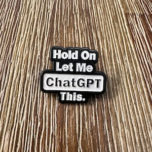 May include: A black and white pin with the text "Hold On Let Me ChatGPT This." The pin has a rectangular shape with rounded corners. The text is in a bold, sans-serif font.