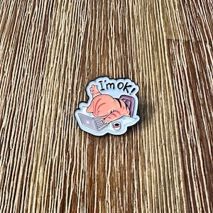 May include: A colorful cat-themed pin with the text "I'm ok!" The pin depicts a cat working on a laptop with a cup of coffee. The cat is orange and white, and the pin has a blue border.