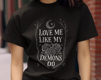 Op de afbeelding: Zwart T-shirt met een gotisch ontwerp. De afbeelding toont de tekst "LOVE ME LIKE MY DEMONS DO" in een gestileerd lettertype, samen met drie rozen en een halve maan. Het ontwerp is omlijst door takken.
