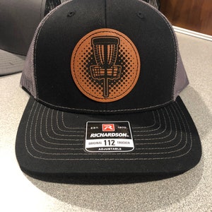 May include: Black and gray trucker hat with a brown leather patch featuring a disc golf basket. The patch has a white outline and a black and white polka dot background. The hat has a black bill and a gray mesh back. The hat has a black label with the text "EST. 1970 RICHARDSON. ORIGINAL 112 TRUCKER ADJUSTABLE".