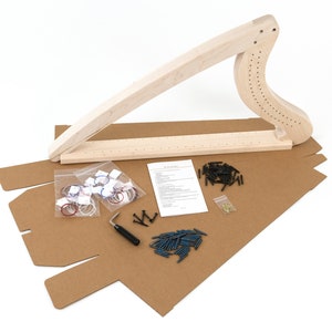 Pode incluir: Um kit de harpa de madeira com todas as peças necessárias para montá-la, incluindo cordas, pinos de afinação e uma ferramenta. O kit está disposto em uma caixa de papelão marrom.