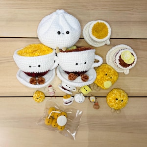 Op de afbeelding: Een verzameling handgemaakte gehaakte voedselartikelen, waaronder een grote witte dumpling, kopjes soep en diverse gebakjes. De items zijn wit, geel en bruin, met schattige geborduurde gezichten. Sommige items staan op schoteltjes.