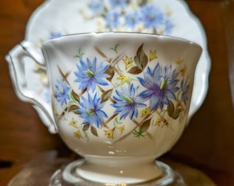 May include: A vintage teacup and saucer set. The cup and saucer are white with a floral pattern of blue and purple flowers, brown stems, and yellow accents. The cup has a clear glass base and a gold rim.
