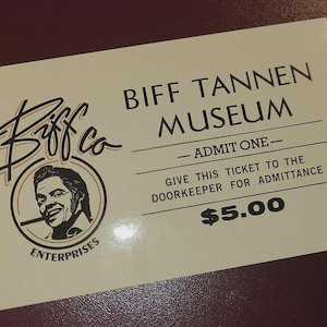 May include: A cream-colored ticket for the Biff Tannen Museum. The ticket features the museum's name, a portrait of a man, and the text "ADMIT ONE". The ticket also states "Give this ticket to the doorkeeper for admittance."